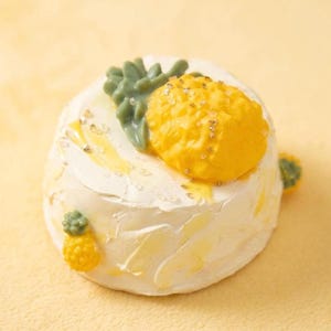 May include: A small, round cake with white frosting, decorated with a yellow pineapple and green leaves. The cake is adorned with gold sprinkles and a miniature pineapple on the side. The background is a solid yellow.