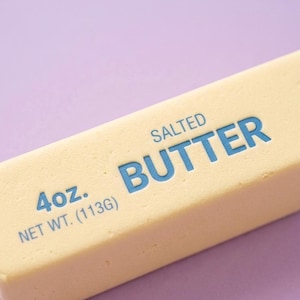May include: A close-up of a stick of salted butter on a purple background. The butter is pale yellow with the word "BUTTER" in blue, along with "4oz." and "NET WT. (113G)" printed on the surface.