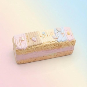 Könnte beinhalten: Ein rechteckiger Biskuitkuchen mit einer hellrosa Cremefüllung. Die Oberseite ist mit vertikalen Streifen aus pastellfarbenem rosa, beigem, gelbem und blauem Zuckerguss verziert, akzentuiert durch kleine, glatte, bunte Bonbons und Streusel.