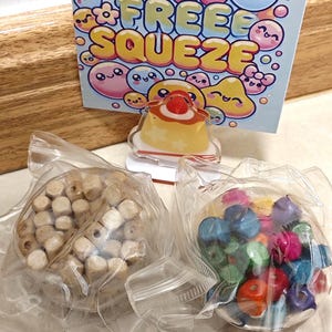 以下が含まれることがあります： 画像には、小さな薄茶色の木製ビーズが入った透明なビニール袋と、カラフルなビーズで満たされたもう1つのビニール袋が2つあります。「STRESS FREE SQUEEZE」と書かれた看板の前には、デザートの小さな透明なアクリルチャームがあります。