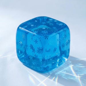 May include: A clear, blue, cube-shaped glass candle holder. The glass is decorated with snowflake designs. The cube is sitting on a white surface, with light reflecting off the glass.