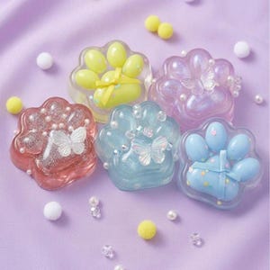 Peut inclure: Cinq ornements en résine en forme de patte de couleur rose, jaune, violet, bleu et bleu clair. Chaque pièce présente des paillettes incrustées, de petites perles blanches et un papillon translucide. Deux pièces ont un ruban noué au centre.