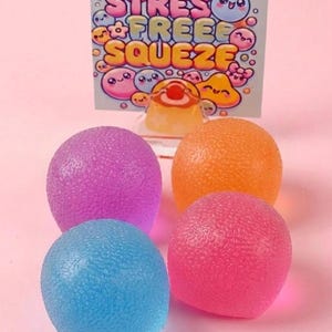 Könnte beinhalten: Vier runde, texturierte Stressbälle in Lila, Orange, Blau und Pink. Hinter den Bällen befindet sich eine rechteckige Karte mit dem Text STRESS FREE SQUEEZE in großen, bunten Buchstaben, umgeben von kleinen, lächelnden Zeichentrickfiguren.