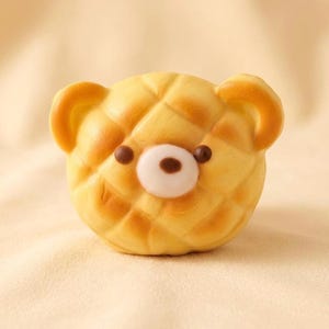 May include: A yellow bear-shaped pastry with a diamond pattern. The bear has brown eyes and a white and brown nose. The ears are a darker shade of yellow. The pastry is on a light yellow background.