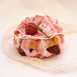 May include: A layered dessert with a light brown base, topped with pink icing and decorative elements. The dessert features a dark brown sphere, a white sphere, and a sprig of greenery. A pink string is tied around the top.