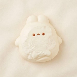 Peut inclure: Une petite figurine blanc cassé aux formes arrondies représentant un lapin stylisé. L'objet présente une surface lisse et mate avec deux petits yeux circulaires brun foncé et une minuscule ligne brune simple formant la bouche au centre.