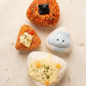 May include: Four triangular onigiri, a Japanese rice ball, are arranged on a textured surface. One is topped with a square of nori seaweed, another with a tomato and sauce, a third with a cartoon character, and the last with cheese and herbs.
