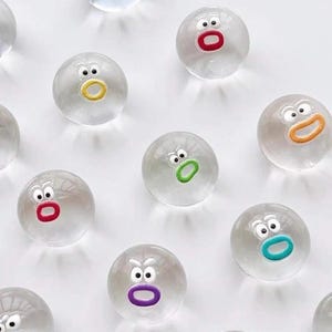 Puede incluir: Una colección de pelotas saltarinas esféricas y transparentes, cada una con un par de ojos blancos y una pequeña boca de forma ovalada en varios colores, incluidos rojo, amarillo, naranja, verde, morado y azul cerceta.