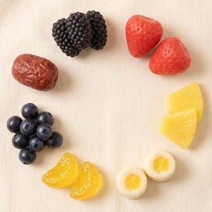 May include: An overhead shot of various fruits arranged in a circular pattern. The fruits include blackberries, strawberries, a date, blueberries, mandarin orange slices, pineapple slices, and two round, white candies with yellow centers. The fruits are arranged on a beige surface.