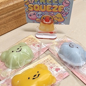 May include: A collection of colorful, squishy stress relief toys. The toys are in the shape of smiling cloud-like figures in green, yellow, and blue, packaged in clear plastic. A small dessert-shaped toy and a sign that reads "STRESS FREE SQUEEZE" are also visible.