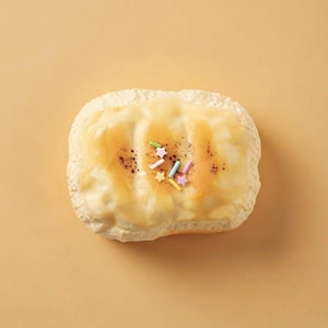 Könnte beinhalten: Ein rechteckiges Gebäck mit einer strukturierten, hellen Kruste, das mit einer glatten, blassgelben Glasur überzogen ist. Die Mitte ist mit einer kleinen Ansammlung bunter, pastellfarbener stern- und zylinderförmiger Streusel sowie feinen braunen Punkten verziert.