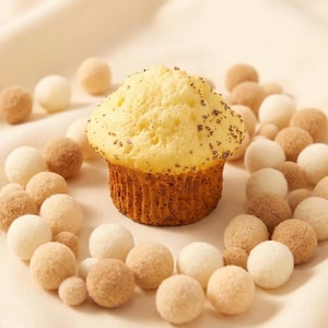 Puede incluir: Un muffin amarillo dorado con una parte superior texturizada y motas oscuras, rodeado de pequeños dulces redondos y de color claro. El muffin se encuentra sobre una superficie clara, creando una disposición visualmente atractiva.