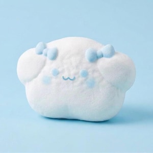 Peut inclure: Une peluche blanche en forme de nuage avec des détails bleu clair. Elle présente deux petits nœuds bleus, des taches bleues sur les joues et un visage souriant. Le fond est bleu clair uni.