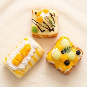 Può includere: Tre pasticcini decorativi in miniatura. Uno ha pezzi di mango e panna montata, un altro una fetta di torta con una foglia verde e una spruzzata di cioccolato, e il terzo ha palline di melone e mirtilli.