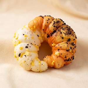 Könnte beinhalten: Nahaufnahme eines croissantförmigen Gebäckstücks. Das Gebäck ist zur Hälfte weiß und zur Hälfte orange, mit schwarzen und gelben Streuseln. Das Gebäck liegt auf einer beigen Oberfläche.