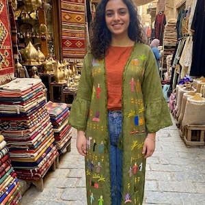 May include: A woman wearing a long, olive green embroidered coat with bell sleeves, over an orange top and blue jeans. The coat features colorful embroidered figures and designs. The background shows a market with textiles and metalwork.