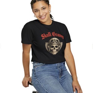 May include: A black short-sleeve t-shirt featuring a graphic of a skull wearing a vintage aviator helmet with goggles and wings. Above the skull, the text "Skull Granny" is printed in a bold red gothic-style font. The shirt is paired with blue jeans.