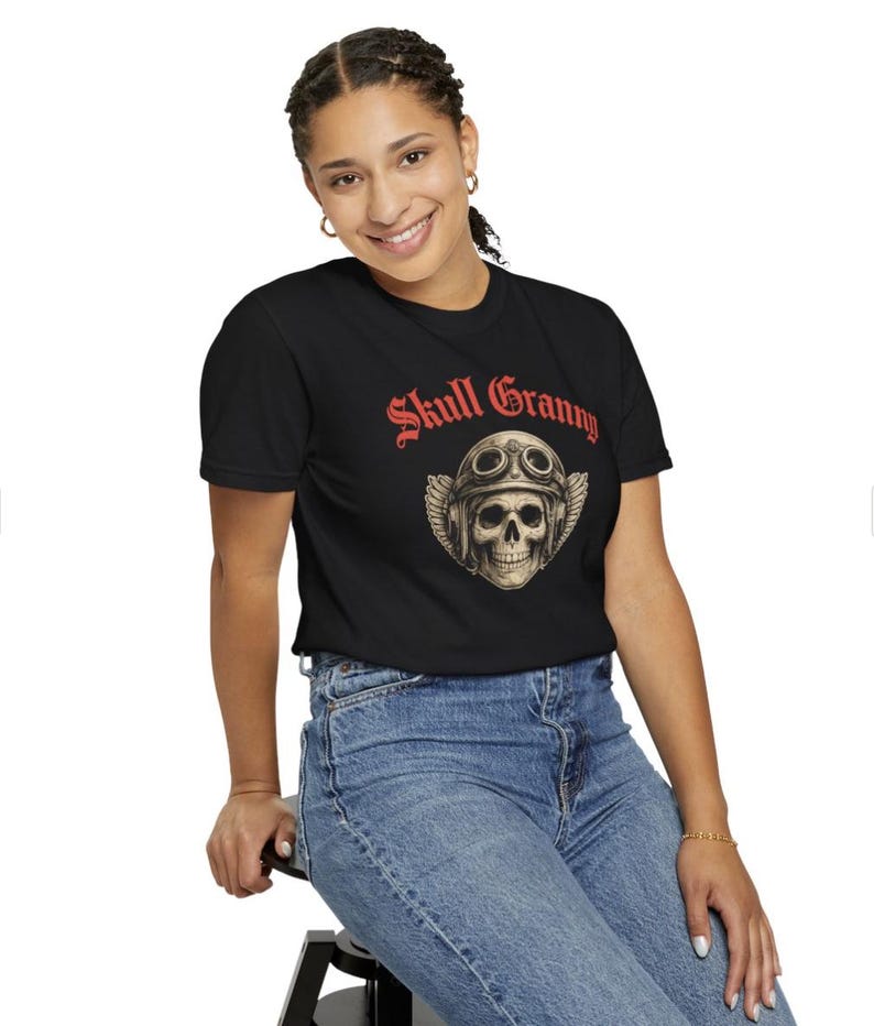 May include: A black short-sleeve t-shirt featuring a graphic of a skull wearing a vintage aviator helmet with goggles and wings. Above the skull, the text "Skull Granny" is printed in a bold red gothic-style font. The shirt is paired with blue jeans.