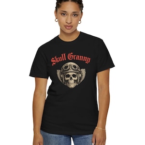 May include: A black short-sleeved t-shirt featuring a graphic of a skull wearing vintage aviator goggles and a leather helmet with wings. Above the skull, the text Skull Granny is printed in a bold, red, gothic-style font on the chest.