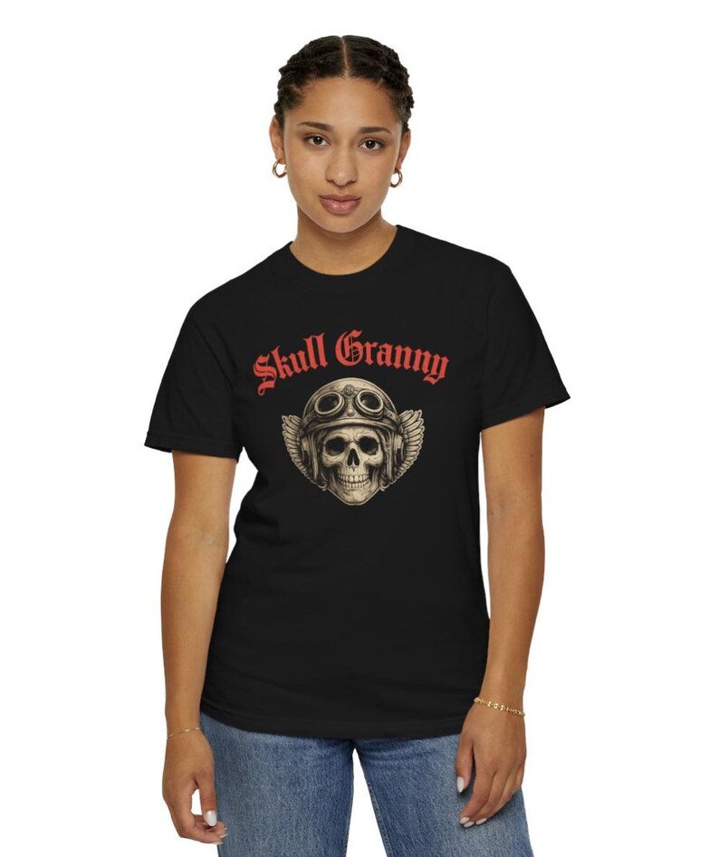 May include: A black short-sleeved t-shirt featuring a graphic of a skull wearing vintage aviator goggles and a leather helmet with wings. Above the skull, the text Skull Granny is printed in a bold, red, gothic-style font on the chest.