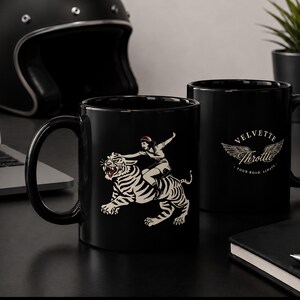 以下が含まれることがあります： 黒いセラミック製のマグカップ2個。片方のマグカップには、赤いアクセントの入った白い虎に乗る人物のグラフィックが描かれています。もう片方のマグカップには、翼のエンブレムの上に白い文字で「VELVETTE Throttle」と書かれ、円状に「YOUR ROAD, ALWAYS」というフレーズが記されています。