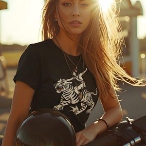 Può includere: T-shirt nero con grafica bianca e nera di una tigre cavalcata da una persona con un cappello rosso. L'immagine è ambientata su uno sfondo luminoso e soleggiato.