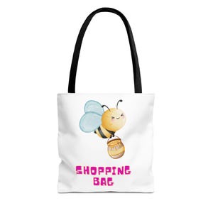 Könnte beinhalten: Eine weiße Tragetasche mit schwarzen Henkeln und einer Grafik einer gelb-schwarzen Biene, die einen braunen Honigtopf trägt. Unter der Biene ist der Text SHOPPING BAG in fetten, pinkfarbenen Großbuchstaben aufgedruckt. Die Tasche ist rechteckig.