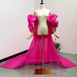 May include: A vibrant hot pink gown featuring voluminous puff sleeves and a long, flowing skirt. The garment is displayed on a mannequin, showcasing a sheer, lightweight fabric with a dramatic train that extends across the floor.