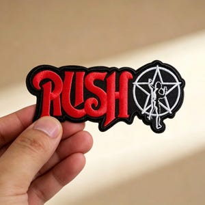 Puede incluir: Parche bordado con la palabra "RUSH" en rojo, letras estilizadas y borde negro. Un diseño de pentagrama blanco y negro con una silueta está a la derecha. El parche está sostenido por una mano.