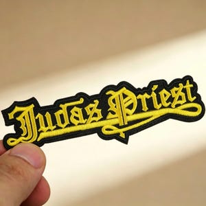 Op de afbeelding: Geborduurde patch met de naam "Judas Priest" in gele letters en gotische letters op een zwarte achtergrond. De patch heeft een zwarte rand en is bedoeld voor kleding of accessoires.