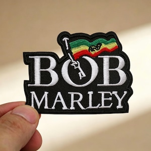 May include: Black embroidered patch featuring the name "BOB MARLEY" in white lettering. A flag with red, yellow, and green stripes is above the name. The patch is held by a hand.