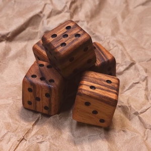 May include: A small stack of five wooden dice with a natural brown wood grain pattern. Each die is cubic in shape with rounded edges and features dark, circular pips representing the numbers one through six on their respective faces.