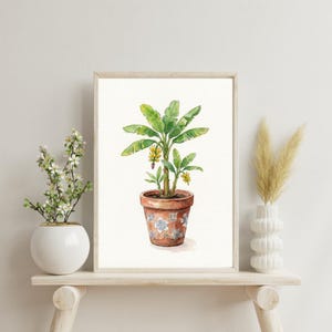 Könnte beinhalten: Aquarellmalerei einer Bananenpflanze in einem Terrakottatopf mit floralem Design. Die Pflanze hat große grüne Blätter und kleine gelbe Blüten. Das Kunstwerk ist gerahmt und wird auf einem Holzregal mit anderen dekorativen Gegenständen ausgestellt.