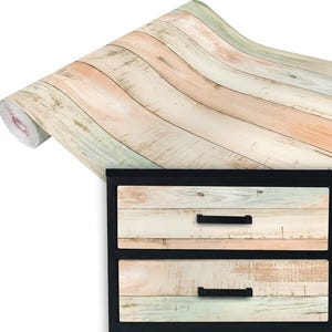 May include: A roll of adhesive contact paper featuring a weathered wood plank pattern in shades of light peach, cream, and pale green. The pattern is shown applied to the two front drawers of a black cabinet with horizontal black metal handles.