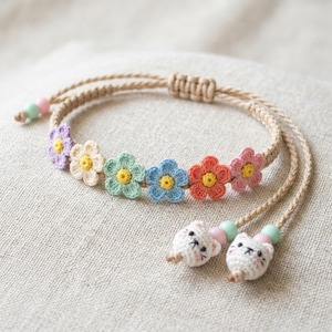 May include: A handmade macrame bracelet featuring a row of colorful crochet flowers in purple, cream, green, blue, orange, and pink. The bracelet has a tan cord and is adorned with two white cat charms with pink and green beads.
