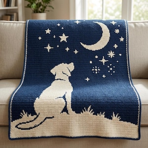 Könnte beinhalten: Eine dunkelblaue Häkeldecke mit der cremefarbenen Silhouette eines Hundes, der auf einem grasbewachsenen Hügel sitzt und zu einer großen Mondsichel und verschiedenen Sternen am Nachthimmel aufblickt, eingefasst mit einem einfachen cremefarbenen Rand.