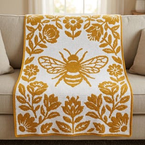 Pode incluir: Uma manta de crochê retangular com fundo branco e um grande motivo central de abelha em amarelo mostarda. O design é emoldurado por uma borda de flores e caules com folhas em amarelo mostarda, criando um padrão floral simétrico no tecido.