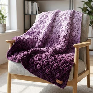 May include: A plush, ombre-dyed blanket in shades of purple, draped over a gray armchair. The blanket features a textured, raised pattern. A small, brown leather tag is sewn on the blanket. The chair has wooden arms and legs.