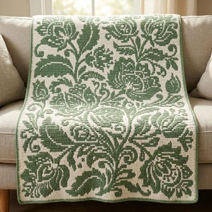 May include: A rectangular crochet blanket featuring a two-tone floral pattern in sage green and cream. The design displays large, intricate blossoms, leaves, and winding stems covering the entire surface in a repeating, symmetrical arrangement.