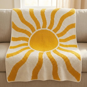 May include: A rectangular crochet blanket featuring a central yellow sun design with radiating yellow sunbeams extending outward against a cream-colored background. The textile is crafted with a consistent stitch pattern throughout the entire piece.