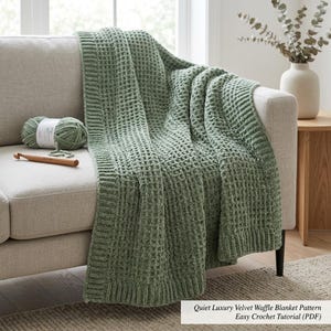 May include: A sage green velvet waffle blanket draped over a light gray sofa. A ball of yarn and a wooden crochet hook rest on the sofa. The blanket has a textured, grid-like pattern. Text on the image reads "Quiet Luxury Velvet Waffle Blanket Pattern Easy Crochet Tutorial (PDF)".