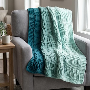 May include: A teal and seafoam green knitted throw blanket draped over a gray armchair. The blanket features a textured, wavy pattern. A small wooden side table with a mug and a plant is visible in the background.