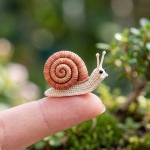 Puede incluir: Un caracol de ganchillo en miniatura con un cuerpo beige y una concha en espiral marrón. El caracol tiene pequeños ojos negros y dos antenas. El caracol descansa sobre la yema de un dedo, mostrando el pequeño tamaño de la artesanía.