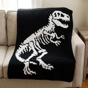 May include: A rectangular crochet blanket featuring a large, white, detailed dinosaur skeleton graphic centred on a solid black background. The design displays the full skeletal structure of a theropod dinosaur in a high-contrast, pixelated style.