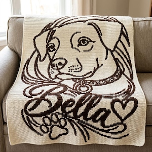 May include: A cream-colored crochet blanket featuring a dark brown graphic design of a dog's face. The design includes the name Bella in cursive script, a paw print, and a heart shape. The blanket is made with a visible yarn stitch pattern.