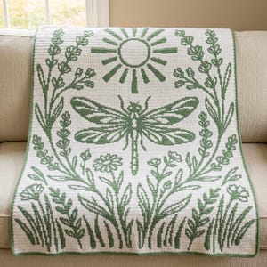 May include: A rectangular crochet blanket featuring a white background with a sage green pattern. The design displays a central dragonfly surrounded by botanical elements, including tall grasses, flowers, and a sun motif at the top center.