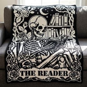 Op de afbeelding: Een zwart-witte gehaakte deken met een skelet dat een boek leest voor een boekenplank. Het ontwerp bevat een maansikkel, rozenranken en een banner aan de onderkant met de tekst THE READER in een vetgedrukt schreeflettertype.