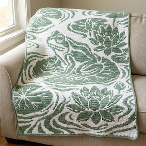Può includere: Una coperta rettangolare all'uncinetto con un motivo bicolore in verde salvia e bianco. Il design mostra una rana centrale seduta su una foglia di ninfea, circondata da ninfee in fiore e increspature stilizzate sull'intera superficie.