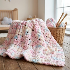 Puede incluir: Una manta tejida en tonos pastel rosa, azul y crema. La manta está hecha de un material suave y afelpado y tiene un diseño de tejido de cesta. Una cesta de hilo y agujas de tejer está cerca. La manta está sobre una superficie de madera.