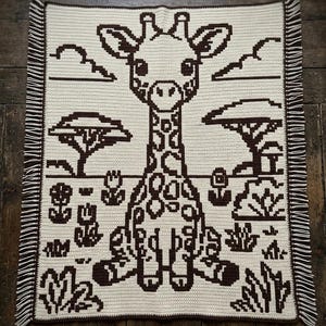May include: A rectangular crochet blanket featuring a dark brown giraffe centred against a cream background. The design includes dark brown silhouettes of acacia trees, clouds, and desert plants. The blanket has a dark brown border and fringe.